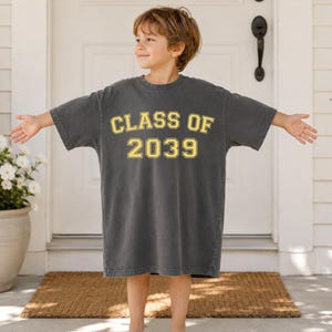 May include: A charcoal gray short-sleeve t-shirt featuring the text "CLASS OF 2039" printed in a yellow collegiate-style font with a black outline. The shirt has a crew neckline and a relaxed, oversized fit that reaches down to the mid-thigh area.