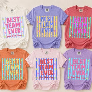 Puede incluir: Seis camisetas de manga corta y cuello redondo en crema, morado, rojo, naranja, rosa claro y lavanda. Cada camiseta presenta un gráfico central con rayas verticales, pequeñas estrellas, el texto "BEST TEAM EVER" y "Your Team Name".