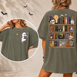 May include: An olive green t-shirt featuring a small graphic on the front of a white ghost reading a book. The back displays a large illustration of wooden bookshelves filled with various books, skulls, a potion bottle, a broom, and a pumpkin.