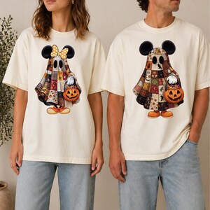 May include: Two cream-colored t-shirts featuring a graphic of a ghost wearing mouse ears and a patchwork-style cloak. The ghost holds a jack-o'-lantern bucket filled with candy. One ghost wears a yellow polka-dot bow, while the other does not.