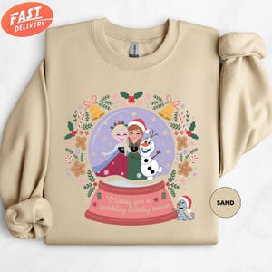 A beige sweater with a Christmas-themed graphic design featuring two girls in a snow globe, surrounded by festive elements such as snowflakes, pine cones, and a dog.