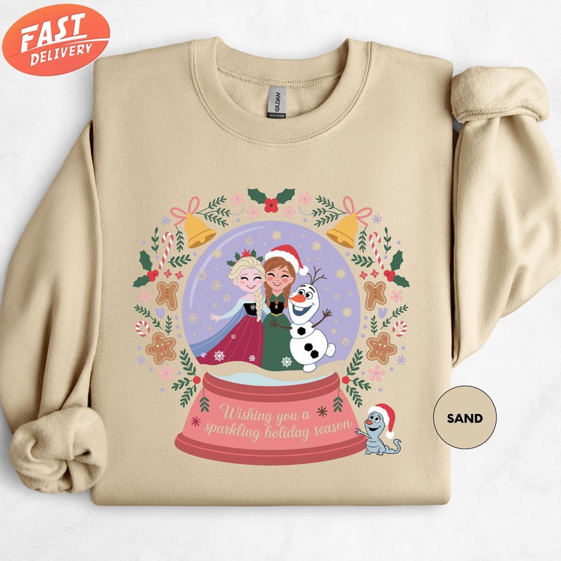 A beige sweater with a Christmas-themed graphic design featuring two girls in a snow globe, surrounded by festive elements such as snowflakes, pine cones, and a dog.