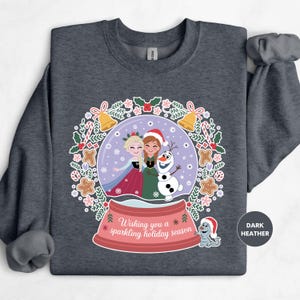 A gray sweatshirt with a graphic design featuring two girls in festive attire inside a snow globe, surrounded by holiday decorations and text wishing the viewer a "sparkling holiday season".