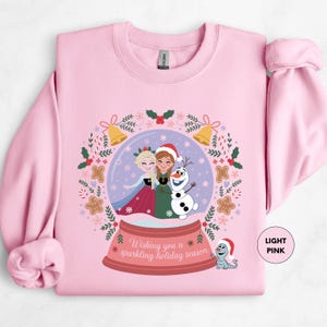 A pink sweatshirt with a graphic design featuring two girls in a snow globe, surrounded by holiday-themed elements such as snowflakes, holly, and a bell.