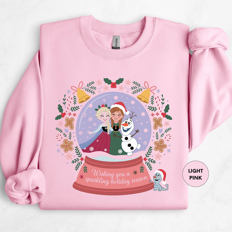 A pink sweatshirt with a graphic design featuring two girls in a snow globe, surrounded by holiday-themed elements such as snowflakes, holly, and a bell.