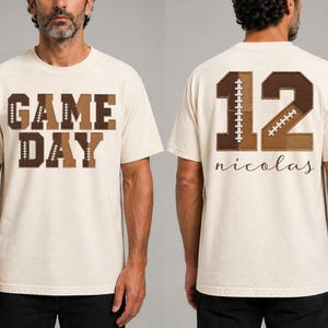 May include: A cream-coloured short-sleeve t-shirt featuring the text GAME DAY on the front in brown block letters with football lace patterns. The back displays the number 12 in brown with football lace details and the name nicolas in script.
