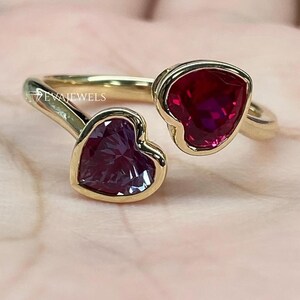 May include: A gold ring with two heart-shaped gemstones. One is a deep red, the other a purplish-red. The ring has a slender band, and the gemstones are set in a gold bezel. The brand name "EVAJEWELS" is visible on the ring.
