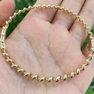 May include: A gold-toned bangle bracelet featuring a continuous twisted, spiral-like design. The circular bracelet has a polished metallic finish and includes a visible hinged clasp mechanism on the side. The text DEVAJEWELS is visible.
