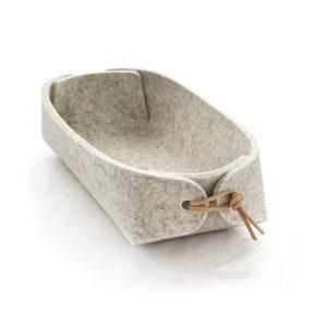 May include: A light gray felt basket with a rectangular shape and rounded corners. The basket has a leather tie closure on each end. The felt material has a textured appearance. The basket is designed for storage.