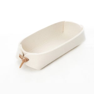 May include: A white felt storage basket with a brown leather handle. The basket is oval shaped and has a slight curve to it.