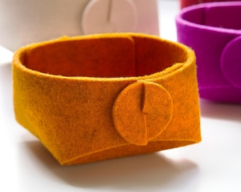 May include: A mustard yellow felt storage basket with a circular button closure on the front. The basket has a structured, open-top design and is made of thick, pressed felt material. Other similar baskets in white, purple, and red are visible.