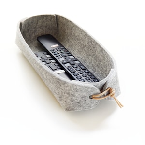 May include: A gray felt storage basket with a leather tie closure, holding two black remote controls. The basket is oval-shaped and sits on a white surface. The remotes have numerous buttons.