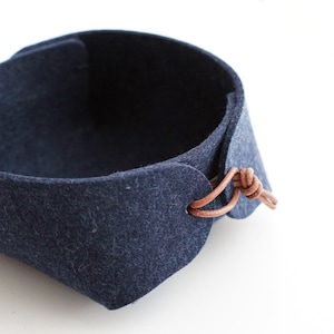 Puede incluir: Un cuenco de fieltro azul marino oscuro con forma cuadrada redondeada. Los bordes del fieltro están unidos en las esquinas con un cordón de cuero marrón claro fino atado en un nudo simple, creando un recipiente de almacenamiento abierto.