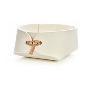 May include: Cream-colored felt basket with a rectangular shape. The basket features a leather tie closure on one side. The design is minimalist and modern, suitable for home organization and storage.