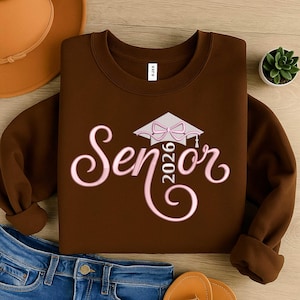 Puede incluir: Una sudadera marrón con la palabra "Senior 2026" en escritura rosa y un diseño de birrete. La sudadera está doblada sobre una superficie de madera con un sombrero, vaqueros y una pequeña planta visibles.