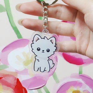 Pode incluir: Um chaveiro de cachorro branco de desenho animado com olhos pretos e contorno preto. O chaveiro é feito de acrílico transparente e está preso a uma corrente e um anel de metal prateado.