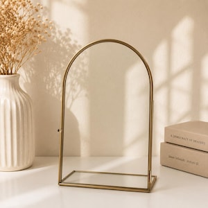 May include: A tabletop mirror featuring a tall, arched glass pane framed in thin, metallic gold-toned metal. The structure includes a rectangular glass base and a hinged design that allows for tilting, providing a clear reflective surface.