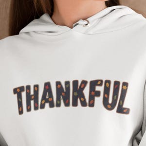 May include: A white hooded sweatshirt featuring the word THANKFUL printed in large, dark charcoal letters. The letters have a stitched-look border and are decorated with a pattern of small, colorful autumn leaves in shades of orange, red, and yellow.