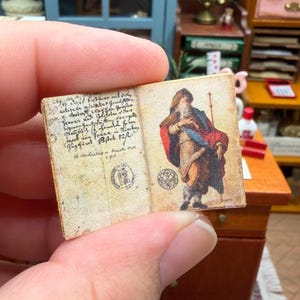 May include: Miniature open book with aged paper pages. The left page has handwritten text and a circular stamp. The right page features a color illustration of a robed figure. The book is held in a hand.