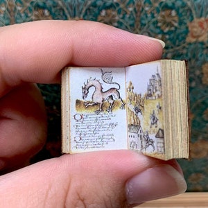 Può includere: Un libro in miniatura con una copertina in pelle marrone è aperto su una pagina con un'illustrazione disegnata a mano di un drago e una città medievale. Il testo sulla pagina è in scrittura corsiva.