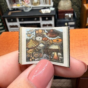May include: A miniature open book featuring a detailed vintage-style illustration of various mushrooms in shades of brown, orange, and white. The text at the top of the page reads Champignons, Fungi, and the book has a dark spine and white pages.