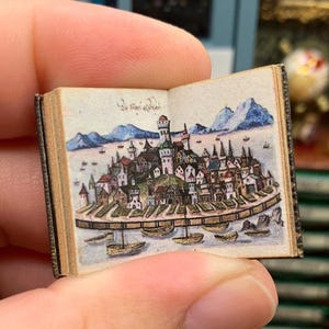 Könnte beinhalten: Ein winziges offenes Buch mit einer detaillierten Illustration einer Küstenstadt. Das Kunstwerk zeigt einen Hafen mit Booten, Gebäuden und Bergen im Hintergrund. Das Buch wird in der Hand einer Person gehalten.