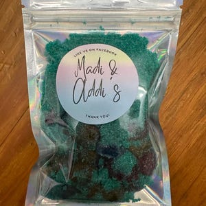 Puede incluir: Una bolsa de plástico transparente llena de exfoliante de azúcar verde. La bolsa tiene una etiqueta circular blanca con el texto "Like us on Facebook Madi & Addi & Thank You!"