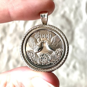 May include: A circular silver-toned pendant featuring a coin design with two embossed doves facing each other above floral details. The year 2000 is stamped at the top center. The pendant is encased in a textured, rope-style metal border frame.