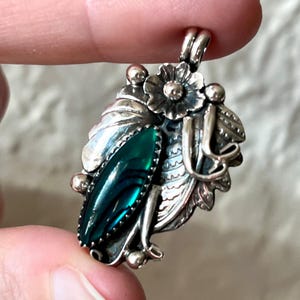 May include: A silver pendant featuring a central marquise-shaped dark green stone with iridescent bands. The metalwork includes detailed leaf motifs, a small flower at the top, and spherical accents. A bail is attached to the top for hanging.