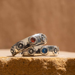 May include: Two silver rings with floral patterns and gemstone details. One ring has a red gemstone, the other a blue gemstone. The rings are presented on a textured surface with the text "Very exquisite".