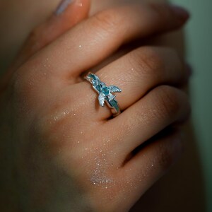 Op de afbeelding: Een zilveren ring met een bloemmotief, bezet met blauwe en heldere edelstenen. De ring wordt aan een vinger gedragen en benadrukt de ingewikkelde details en sprankelende accenten. Het ontwerp van de ring is delicaat en elegant.