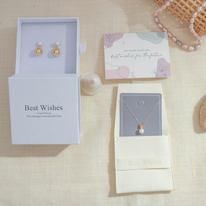 May include: A jewelry set featuring gold-toned earrings and a necklace with a pearl pendant. The earrings are displayed in an open white box with the text "Best Wishes." A card with the text "best wishes for the future" is also present.
