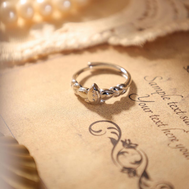May include: A silver-colored ring with a teardrop-shaped centerpiece adorned with three small, clear stones. The ring features a delicate, adjustable band and is set against a backdrop of vintage paper with script and floral designs. The ring is a piece of jewelry.