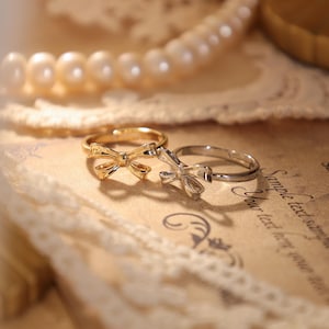 Può includere: Due anelli con design a forma di fiocco. Uno è dorato, l'altro argentato. Gli anelli sono su uno sfondo in stile vintage con una collana di perle e pizzo.