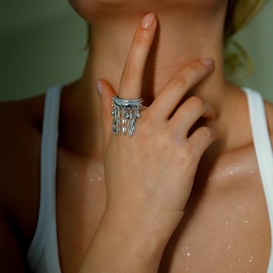 May include: A silver ring featuring multiple bands and dangling geometric charms. The ring is worn on a finger, with the hand and arm visible. The ring's design is intricate, with a modern and artistic aesthetic. The background is a soft, neutral colour.