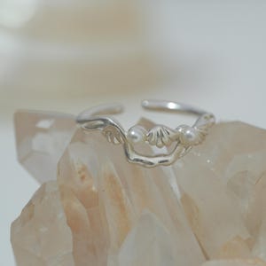 Puede incluir: Anillo plateado con diseño de concha y dos pequeñas perlas. El anillo tiene un diseño abierto, lo que permite ajustar la talla. Se muestra sobre una superficie clara y texturizada.