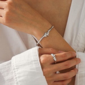 Könnte beinhalten: Ein silberfarbenes Armband- und Ring-Set. Das Armband ist ein schmaler Band mit einem Blumendesign in der Mitte. Der Ring hat ein passendes Blumendesign. Beide werden am Arm und an der Hand einer Person vor einem weißen Hemd getragen.