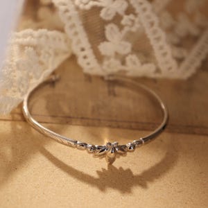 May include: A silver-toned bracelet with a bee-shaped centerpiece. The bracelet has a delicate design with small decorative elements along the band. The background includes lace and a vintage paper.