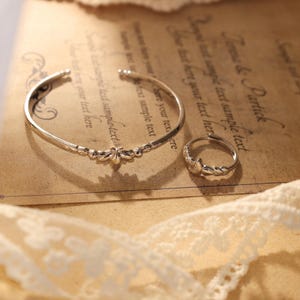 May include: A silver-toned bracelet and ring set. The bracelet is a cuff style with a floral design in the center. The ring has a matching floral design. Both pieces are on a vintage paper background with lace trim.