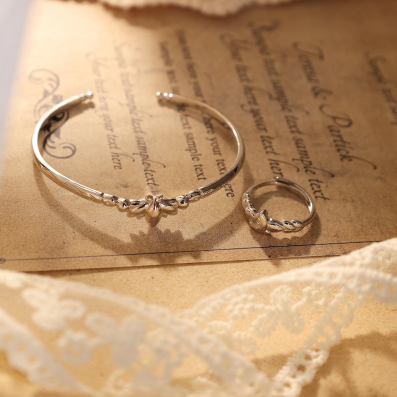 May include: A silver-toned bracelet and ring set. The bracelet is a cuff style with a floral design in the center. The ring has a matching floral design. Both pieces are on a vintage paper background with lace trim.