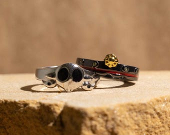 Könnte beinhalten: Zwei Ringe werden gezeigt. Einer ist silberfarben mit einem Totenkopf-Design und schwarzen Augen. Der andere ist schwarz mit goldenem Akzent und rotem Streifen. Die Ringe liegen auf einer strukturierten, hellbraunen Oberfläche.