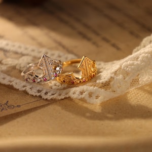May include: Two crown-shaped rings, one silver and one gold, rest on a lace-trimmed surface. The silver ring features small red gemstones and intricate detailing. The gold ring also has red gemstones and a detailed design, showcasing a regal aesthetic.