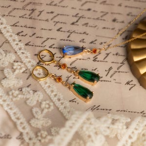 Op de afbeelding: Een goudkleurige sieradenset met een ketting en oorbellen. De ketting heeft een blauwe druppelvormige hanger met oranje accenten. De oorbellen hebben groene druppelvormige edelstenen en oranje accenten. De sieraden staan op een vintage papieren achtergrond.