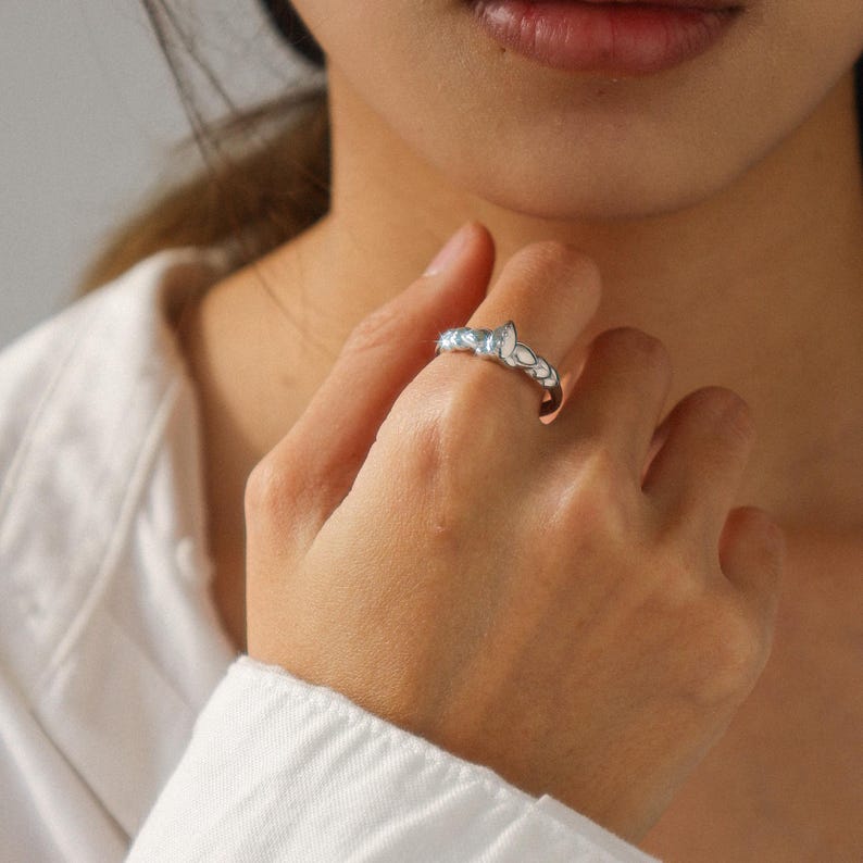 May include: A silver ring with a delicate floral design, featuring small, light blue accents. The ring is worn on a finger, showcasing its intricate details and elegant style. The ring's design includes a series of small, petal-like shapes.