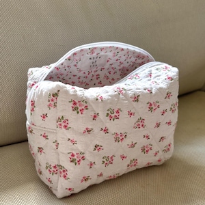 May include: White quilted cosmetic bag with a floral pattern of pink roses and green leaves. The bag has a zipper closure and a matching floral interior. The bag is sitting on a beige surface.