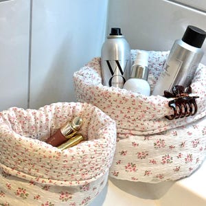 Peut inclure: Deux paniers de rangement en tissu à motifs floraux. Les paniers sont blancs avec de petites fleurs roses. L'un contient du maquillage, l'autre des produits capillaires et de soin. Les paniers sont sur une surface blanche.