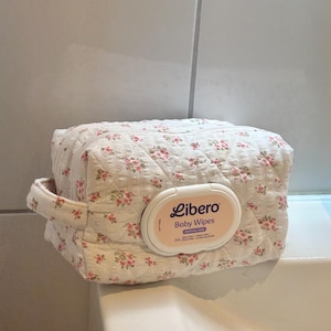 Könnte beinhalten: Eine rechteckige, gesteppte Stofftasche mit cremefarbener Basis und kleinem rosa Blumenmuster. Die Tasche verfügt über einen seitlichen Griff und einen weißen Kunststoff-Spenderdeckel mit der Aufschrift Libero Baby Wipes, Perfume Free, 64.