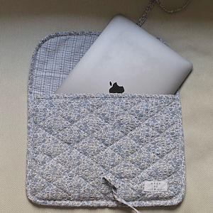 May include: A light blue quilted laptop sleeve with a floral pattern. The sleeve is open, revealing a silver laptop. The interior lining is a light blue and white checkered pattern. The sleeve has a tie closure and a small tag that reads "made by [illegible]"