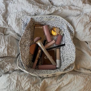 May include: A fabric storage basket with a white background and a small blue floral pattern, filled with various makeup products including a large powder brush, several lipstick tubes, a concealer wand, and compacts in gold and brown tones.