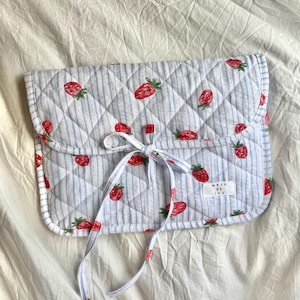 Könnte beinhalten: Gesteppte Clutch mit blau-weißem Streifenmuster, verziert mit roten Erdbeer-Illustrationen. Die Clutch hat einen Schleifenverschluss und ein kleines Etikett mit der Aufschrift "made by lou".